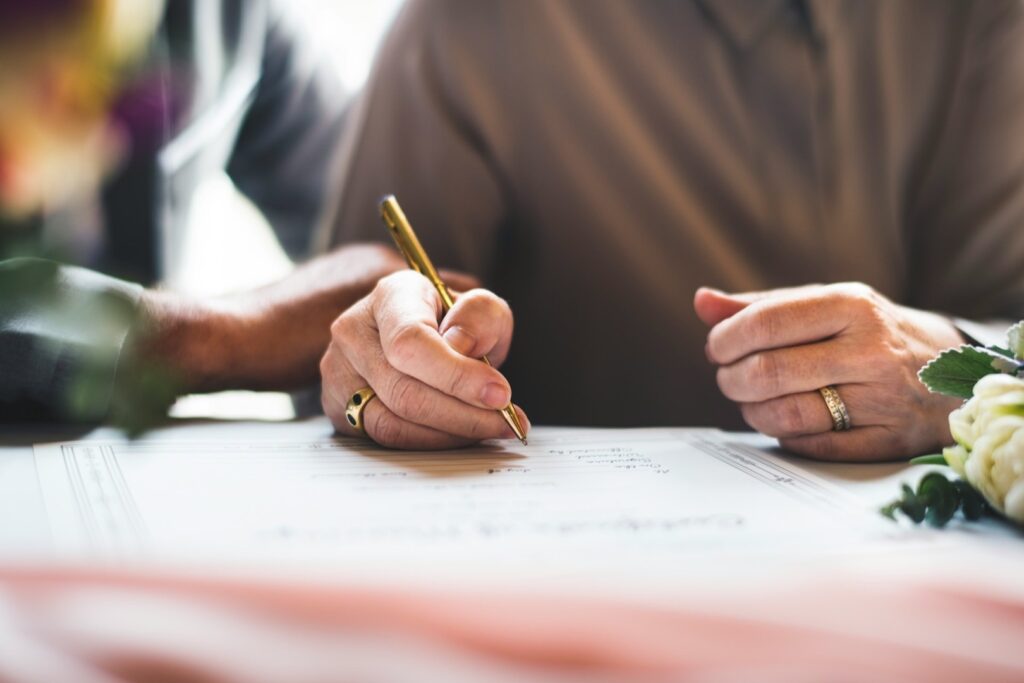 Hand wearing a wedding ring signs a document with a gold pen beside white flowers